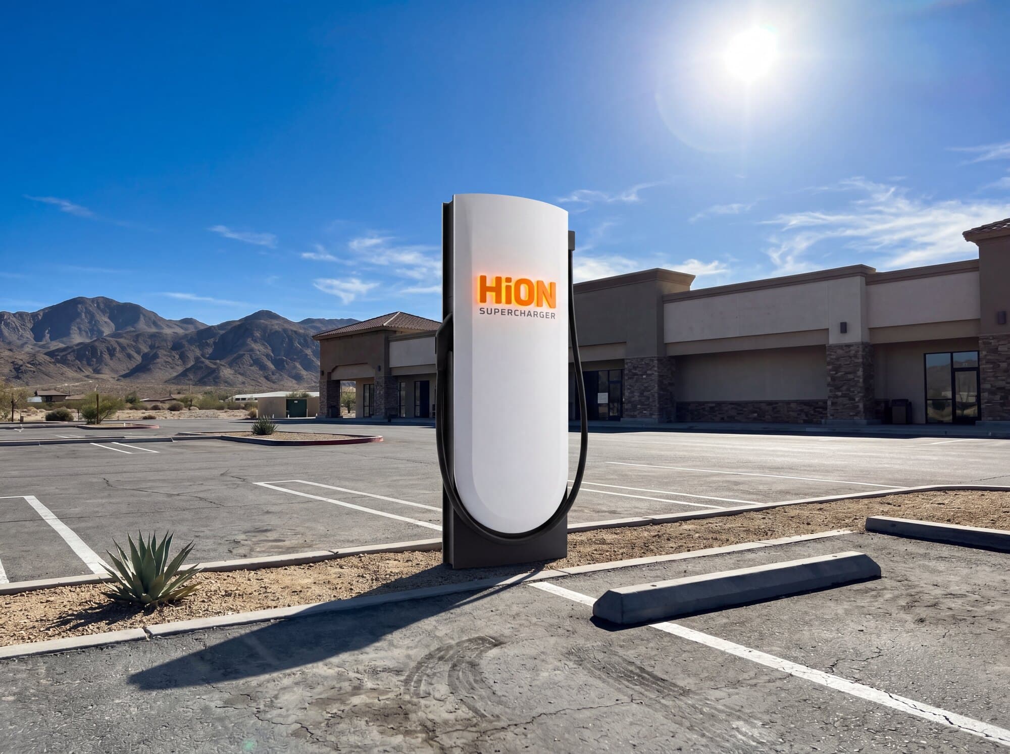 A HiON Supercharger pillar at a sunlit desert roadside stop, a low retail building and open parking behind it, the matte-white face marked with the glowing orange HiON wordmark.